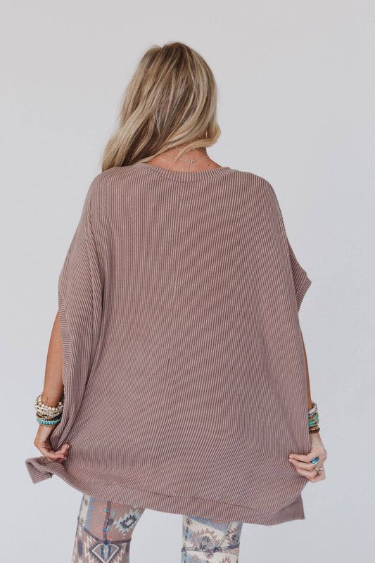 A female model wearing an oversized knit top in brown color, paired with patterned leggings, accessorized with a necklace and multiple bracelets.