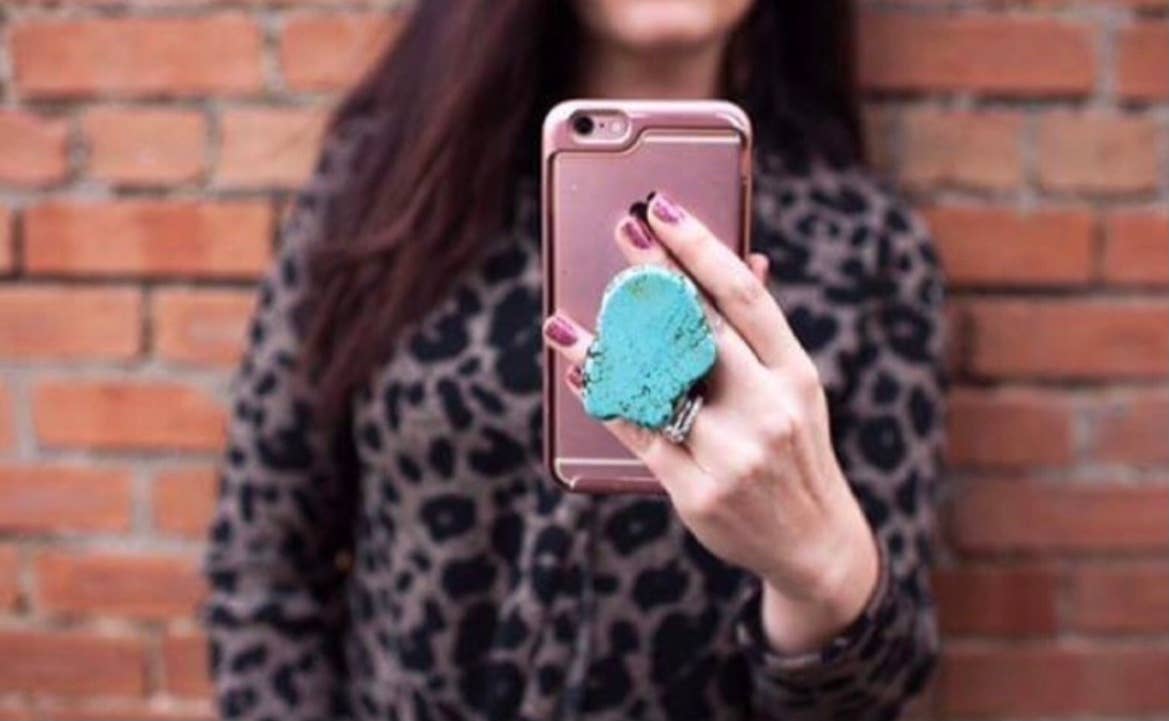 A person holding a smartphone with a turquoise phone grip attached to it, in front of a brick wall.