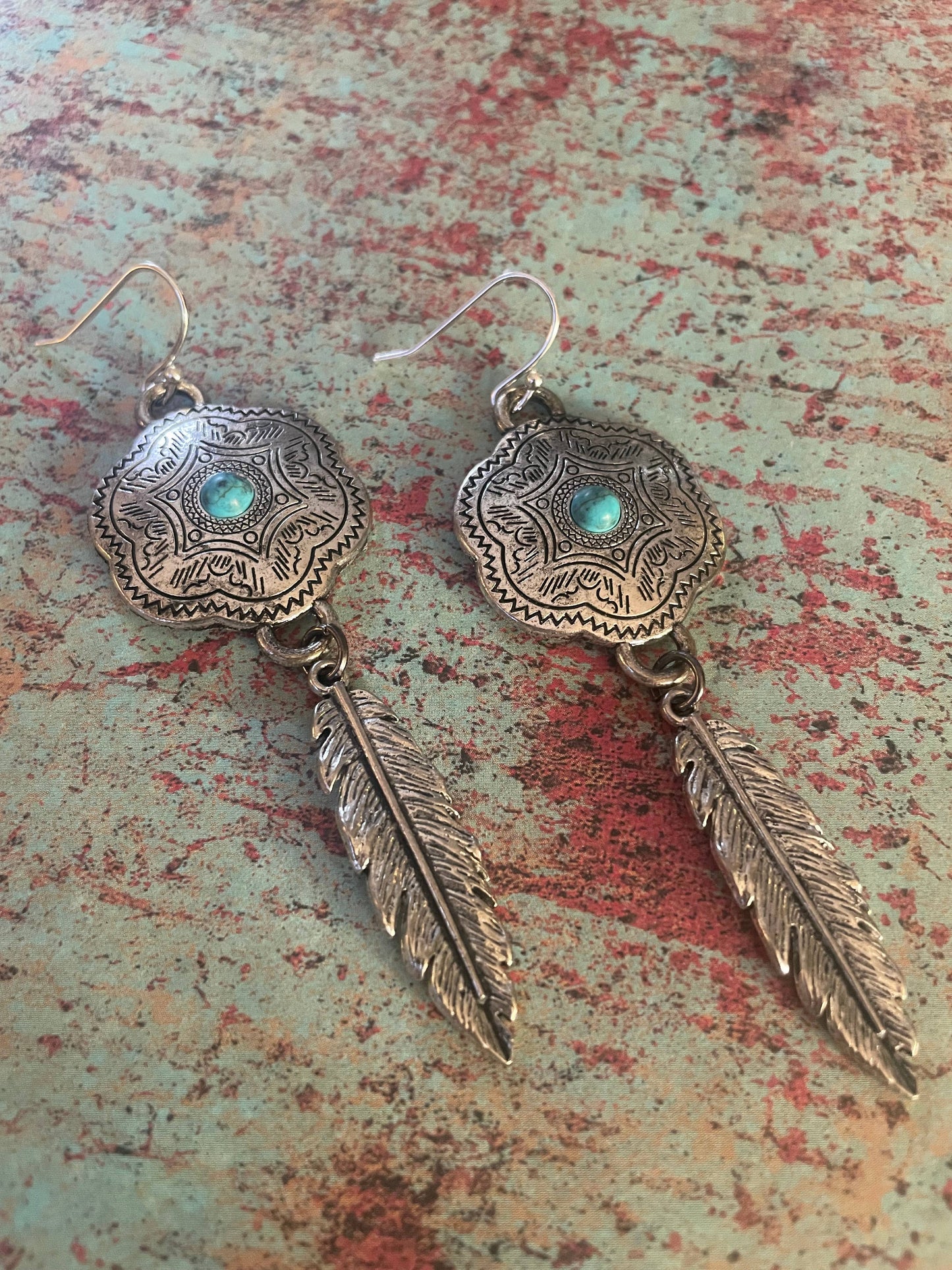 A pair of silver-tone concho earrings with feather charm dangles on a textured background.