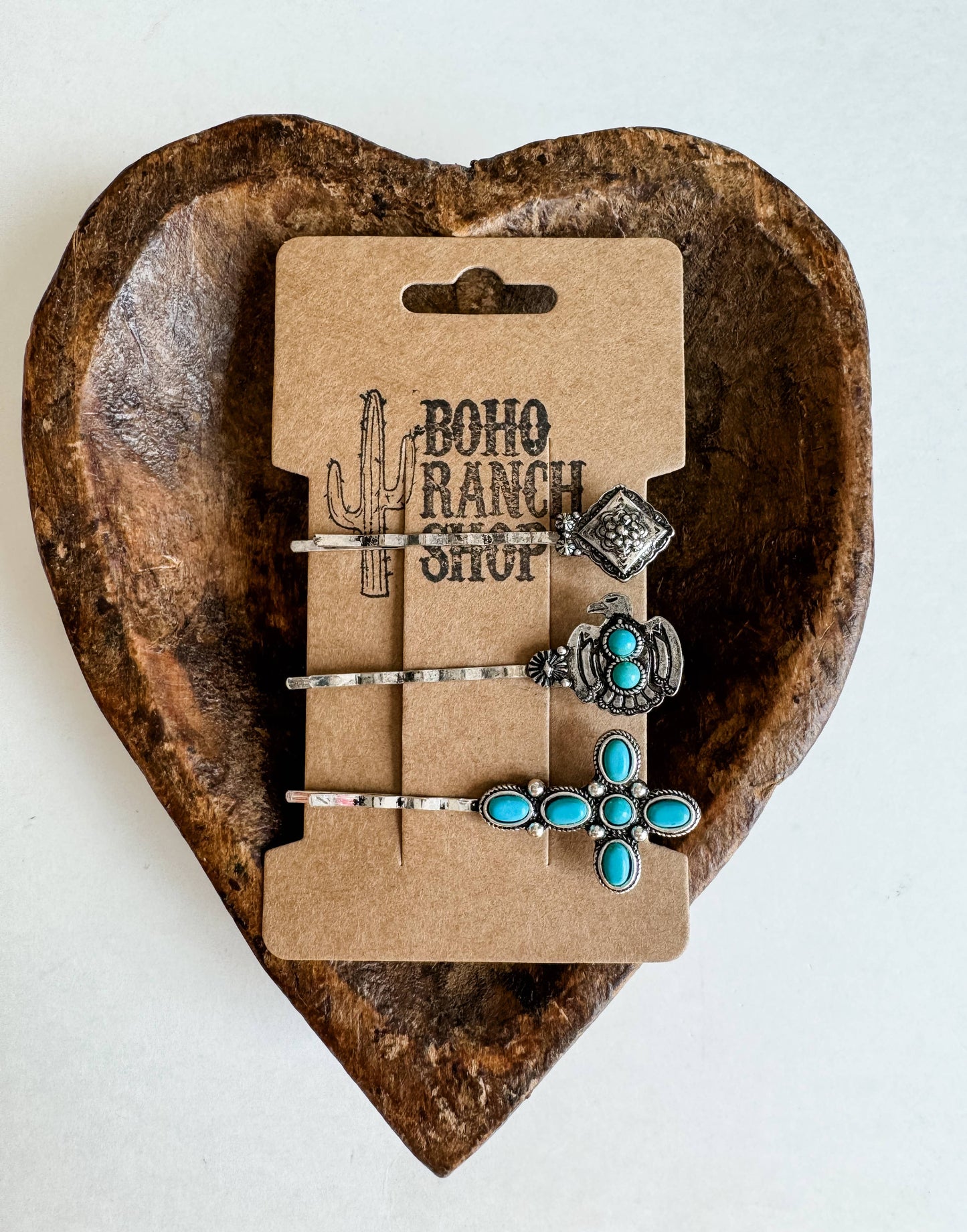 A set of three hair bobby pins with silver and blue floral designs, displayed on a cardboard backing with 'BOHO RANCH SHOP' written on it, placed on a rustic brown wooden surface.
