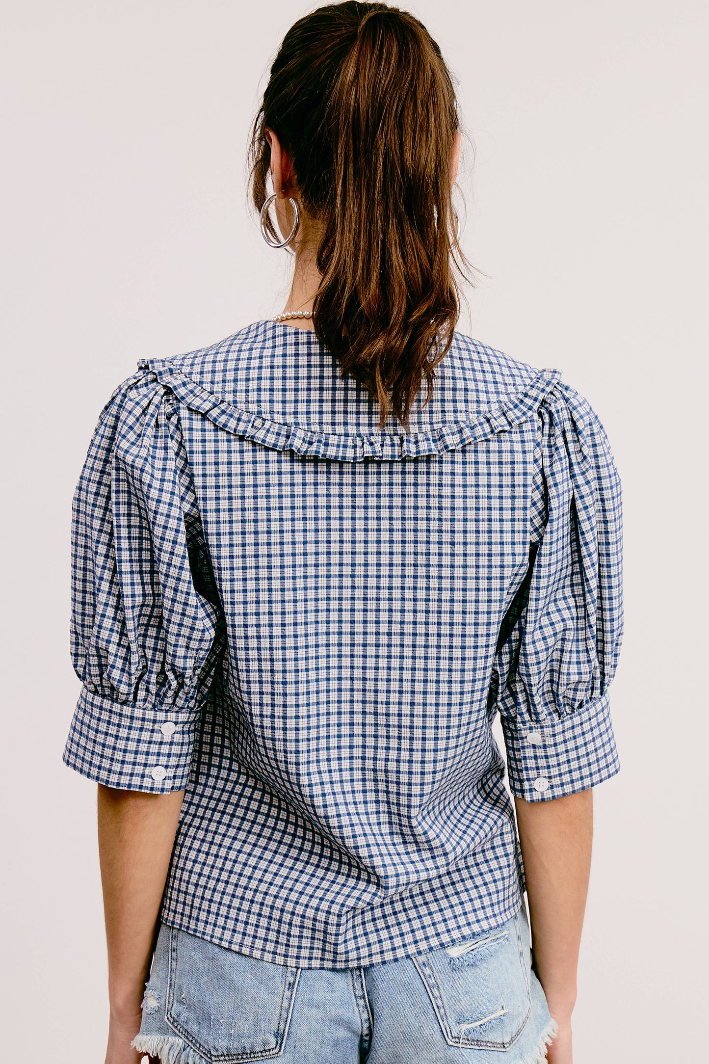 A woman wearing a blue and white gingham print blouse with a round neckline, short puffed sleeves, and a tie at the chest. She is also wearing denim shorts and a brown belt.
