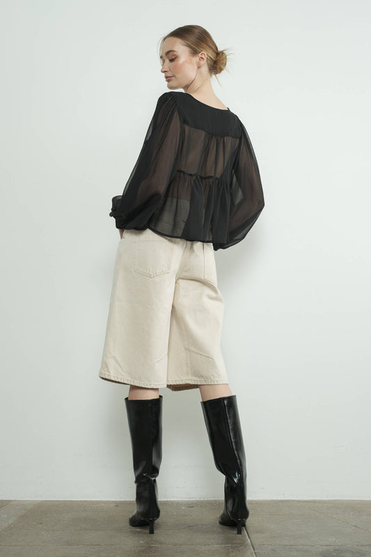 Woman wearing a black sheer top, beige shorts, and black boots against a white background