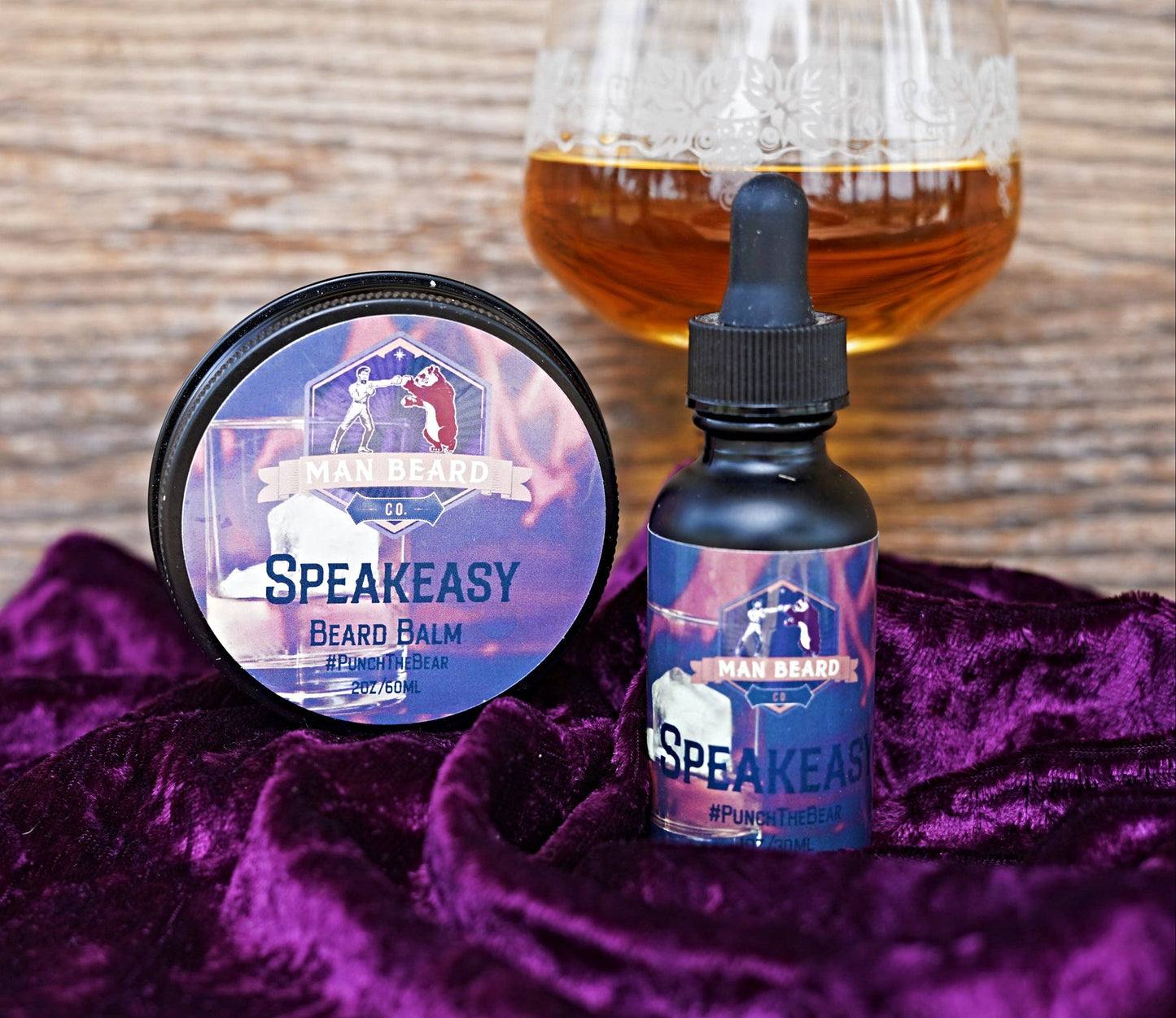 A glass with ice and a beard oil product inside, with the word 'Speakeasy' written on it, set against a backdrop with a warm, fiery appearance.