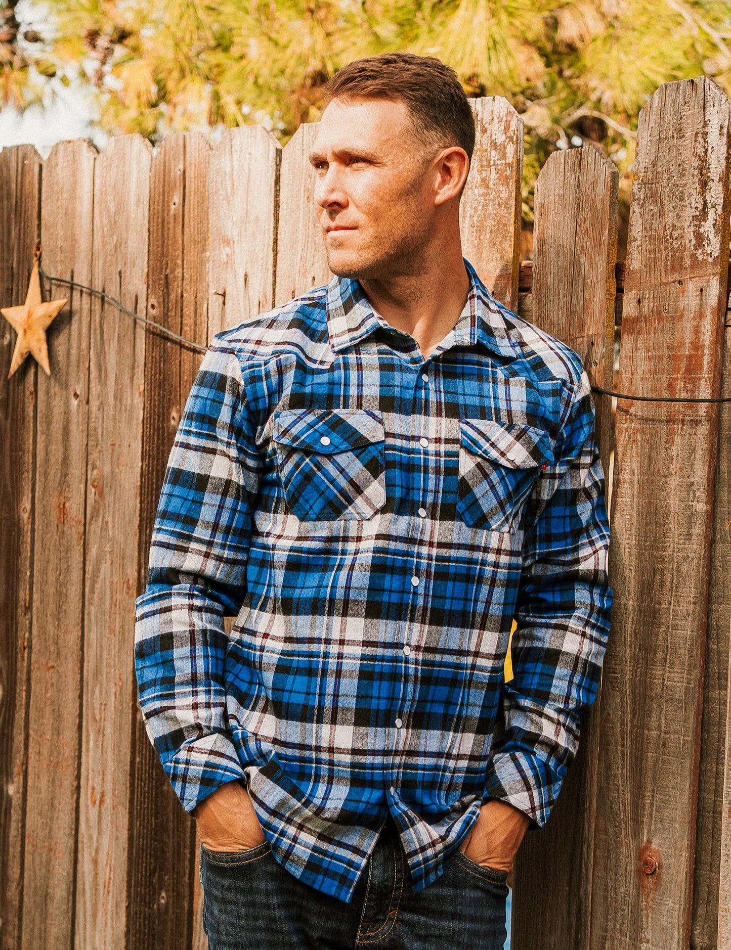 A person wearing a blue checkered long-sleeved shirt standing by a wooden fence.