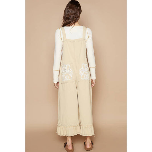 A female model wearing beige overalls with ruffle detail and back pockets, paired with a white top and black sandals, standing against a beige background.