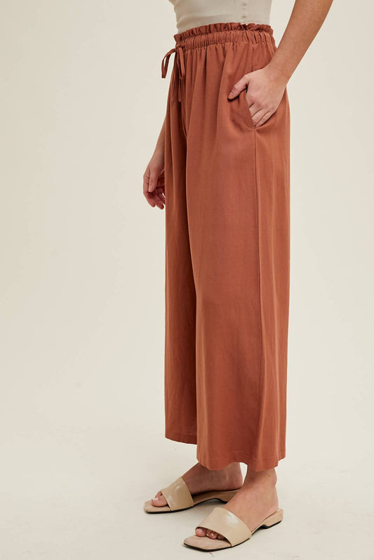 A person standing sideways, wearing wide leg linen pants in a brick color and a matching top, with their hands in the pockets.