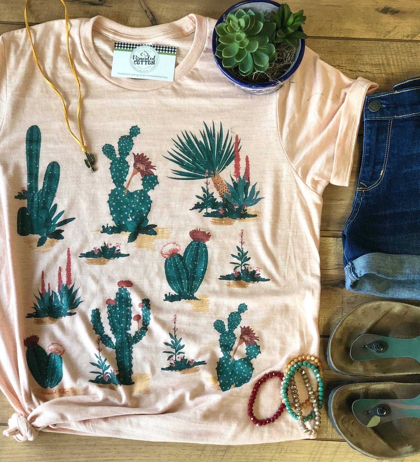 A peach-colored t-shirt with a graphic print of various cacti, displayed flat with a small bowl of cactus, a pair of denim shorts, and some accessories like a watch and a bracelet.