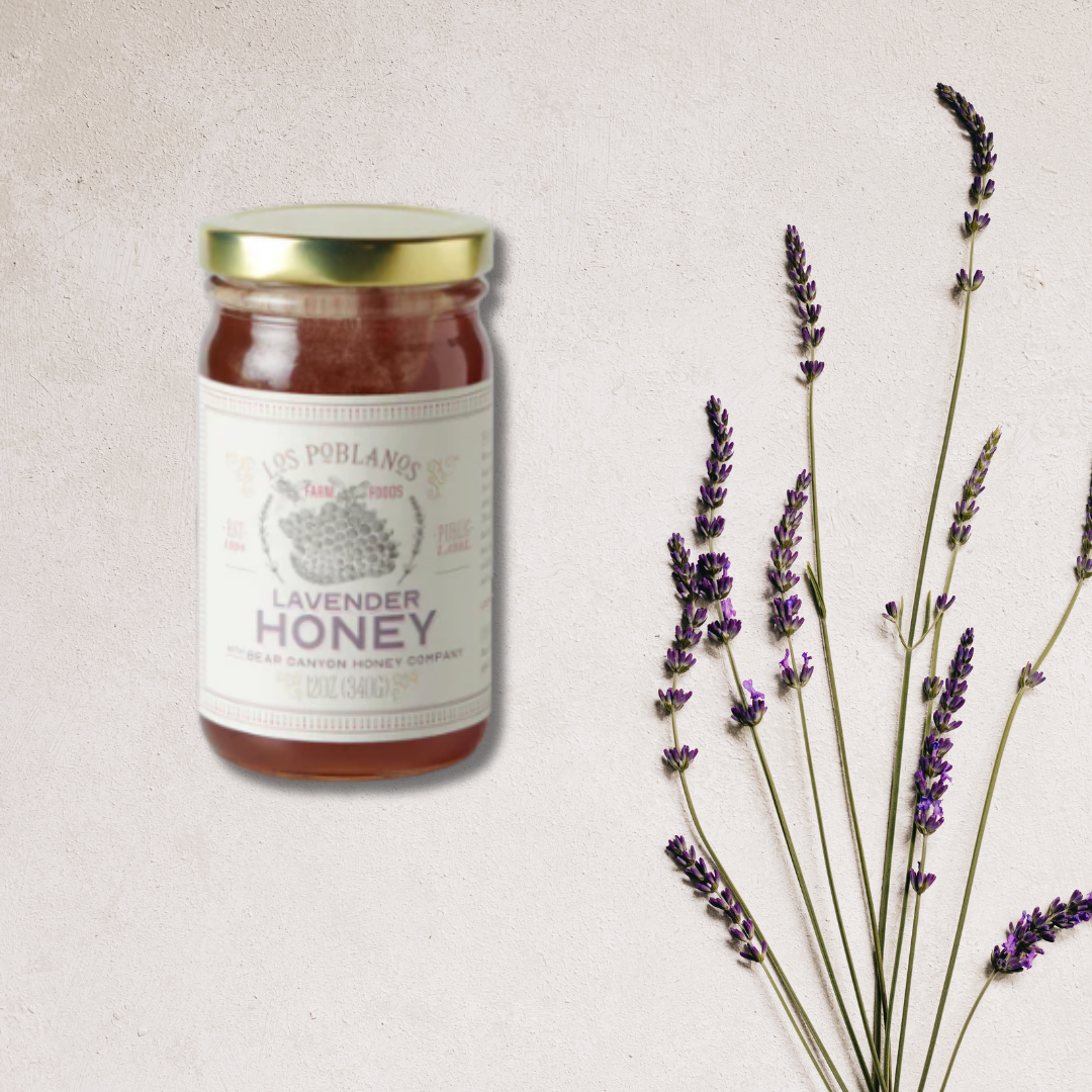 A jar of Lavender Honey labeled Los Poblanos with a background of light-colored wall and a bundle of lavender sprigs to the right of the jar.