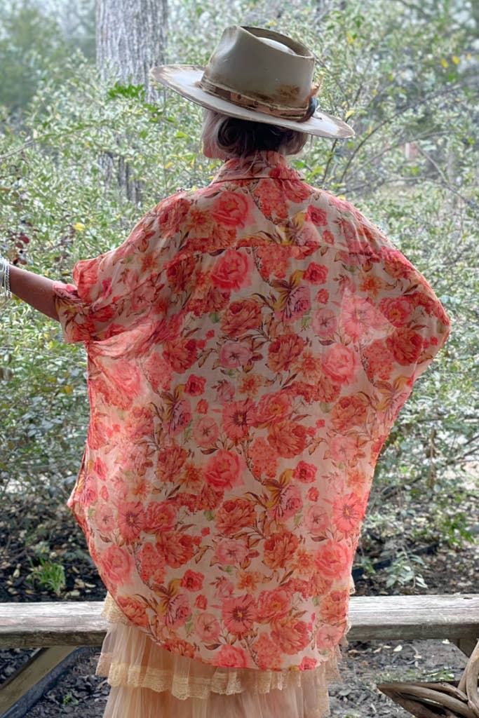 A person wearing a floral top with orange, pink, and red colors, paired with a beige skirt and accessories, standing outdoors.