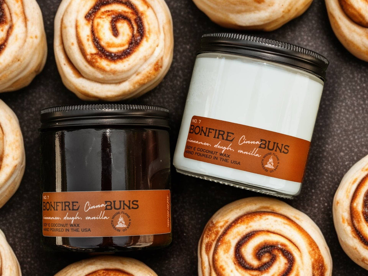 Candle jars labeled 'Bonfire Cinnamon Buns' surrounded by cinnamon rolls on a dark surface.