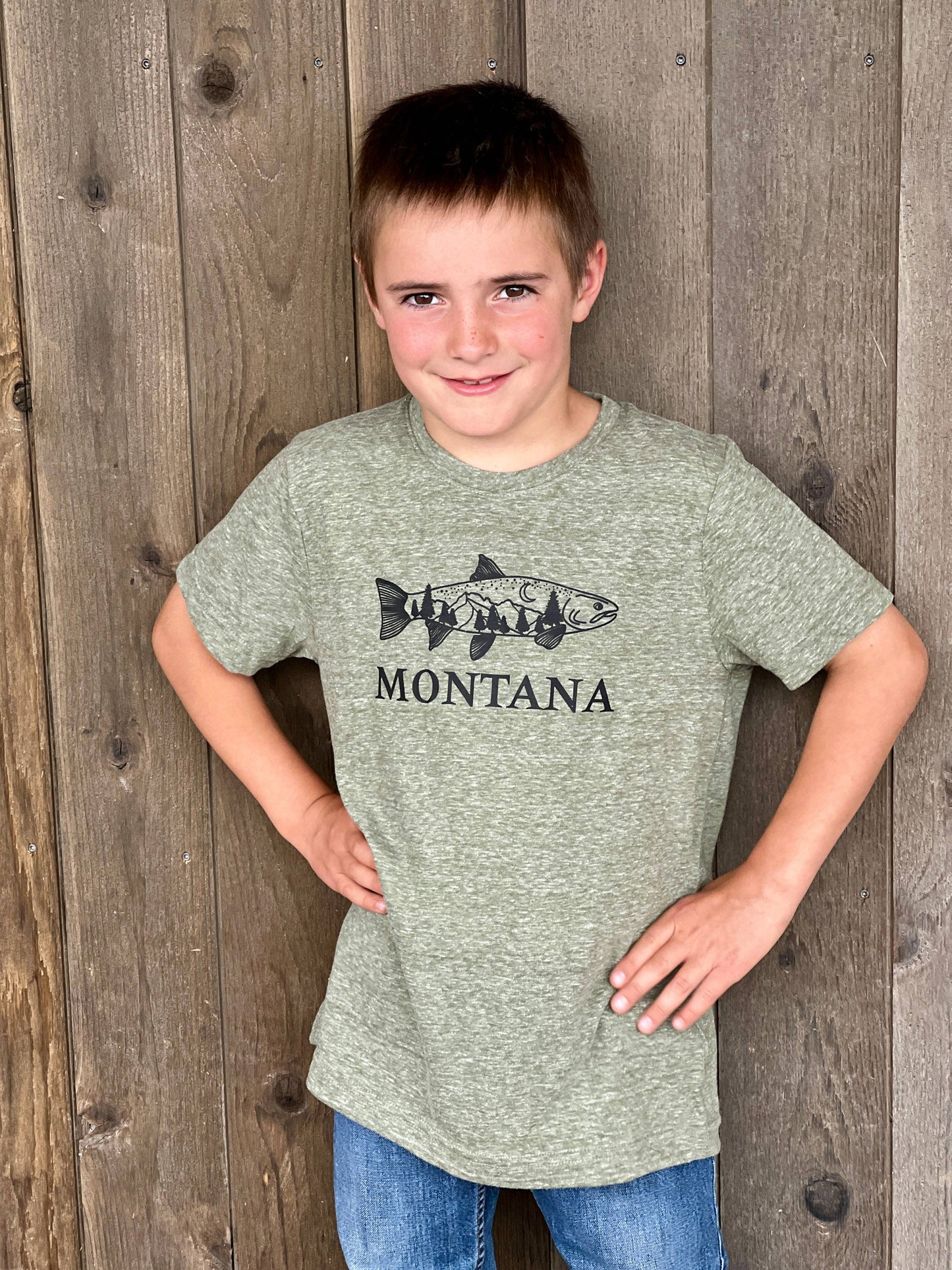 A child standing in front of a wooden backdrop wearing a green t-shirt with 'MONTANA' and a fish graphic on it.