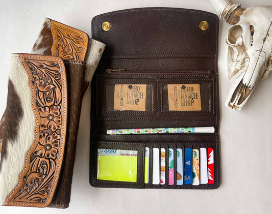 A leather cowhide wallet with intricate tooling, displayed next to a passport, boarding pass, and an inflight entertainment guide on an airplane seat.