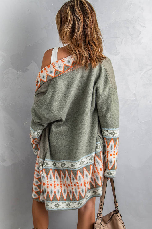 A woman wearing an open front knitted cardigan with geometric patterns in beige, gray, and orange colors over a white top and blue denim shorts.