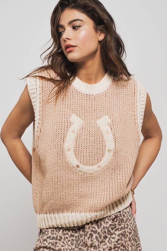 A female model wearing a beige short sleeve sweater vest with a grey embroidered horseshoe on the front, paired with a leopard print skirt.