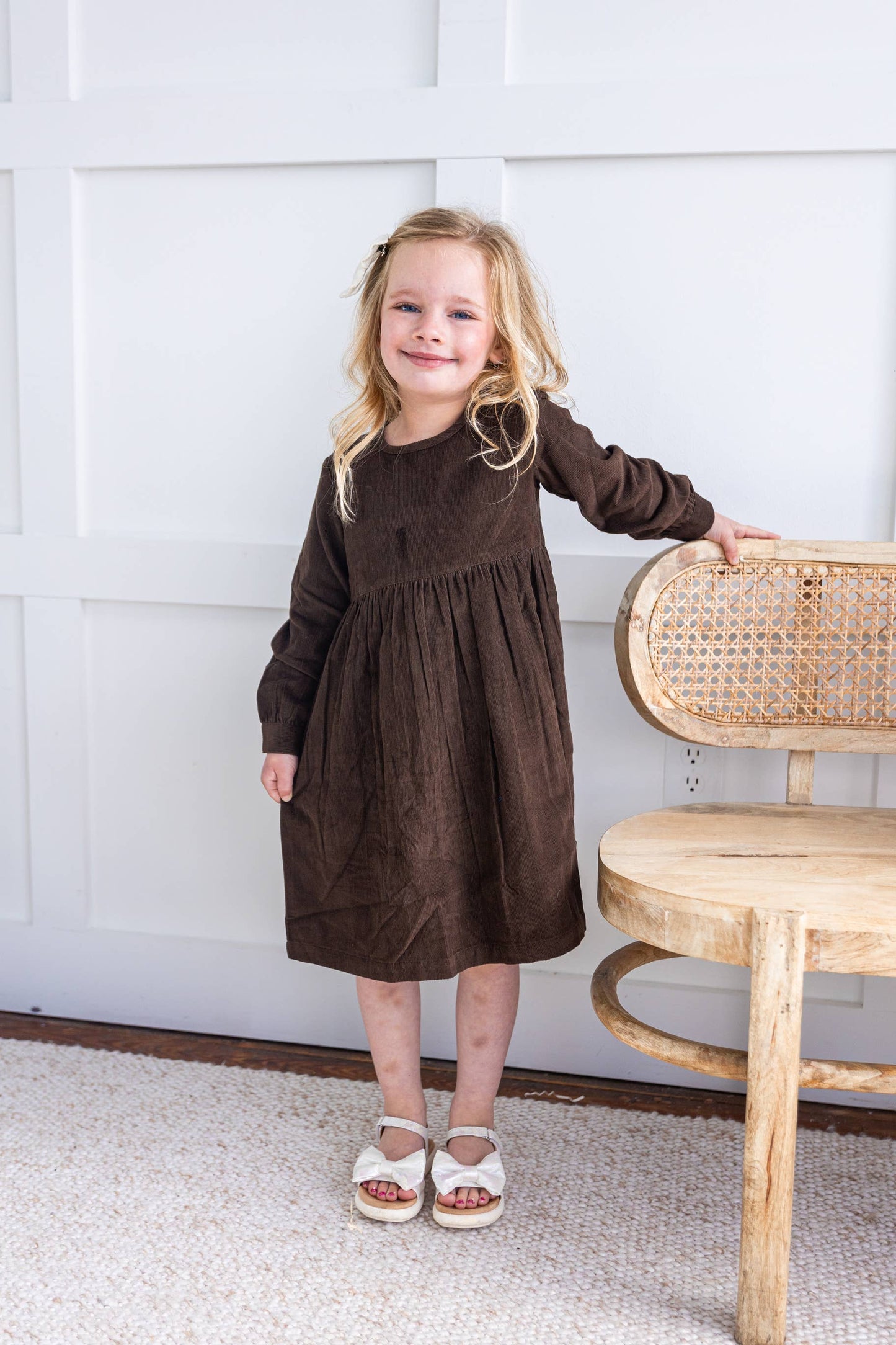 A child standing next to a chair wearing a brown corduroy dress with long sleeves and knee-length.