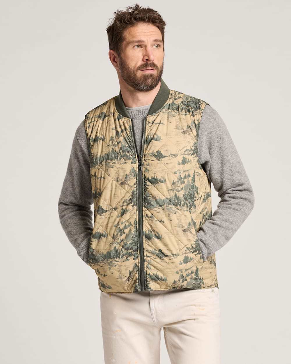 Man wearing a camouflage-patterned vest over a gray long-sleeve shirt on a light gray background