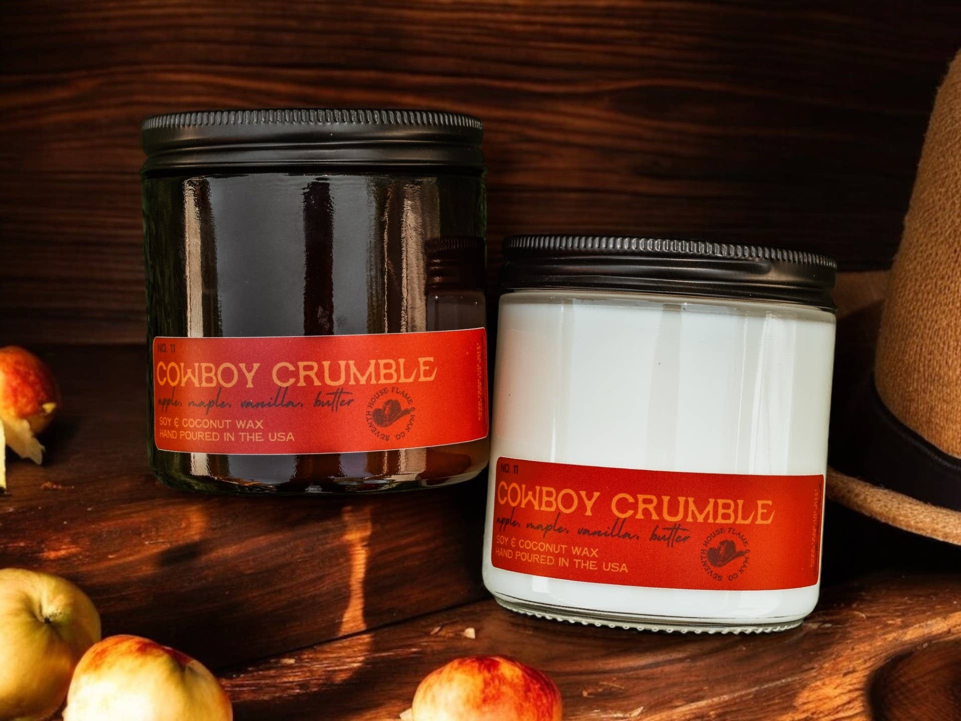 Two 'Cowboy Crumble' candles on a wooden surface with apples and a hat in the background.