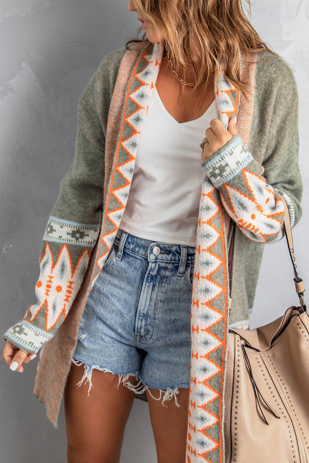 A woman wearing an open front knitted cardigan with geometric patterns in beige, gray, and orange colors over a white top and blue denim shorts.