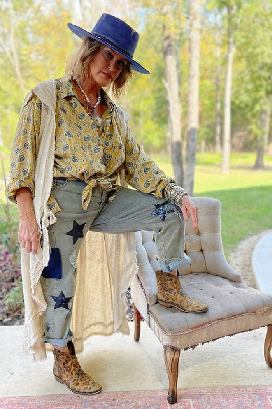 A person seated on a chair wearing a mustard paisley patterned top with star appliques, long pants, boots, and a wide-brimmed hat.