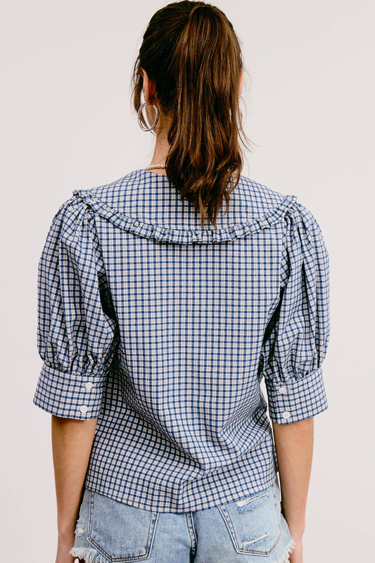 A woman wearing a blue and white gingham print blouse with a round neckline, short puffed sleeves, and a tie at the chest. She is also wearing denim shorts and a brown belt.