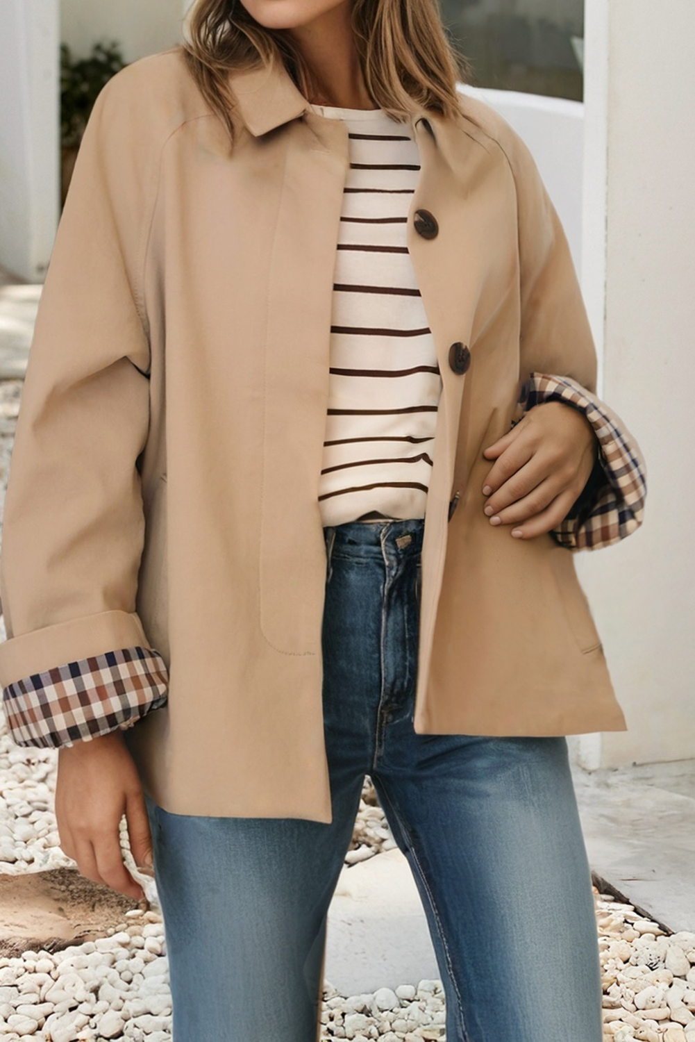 Person wearing a beige trench coat over a striped shirt with plaid cuffs, standing outdoors.