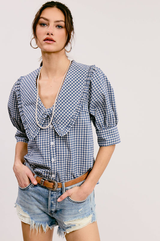 A woman wearing a blue and white gingham print blouse with a round neckline, short puffed sleeves, and a tie at the chest. She is also wearing denim shorts and a brown belt.