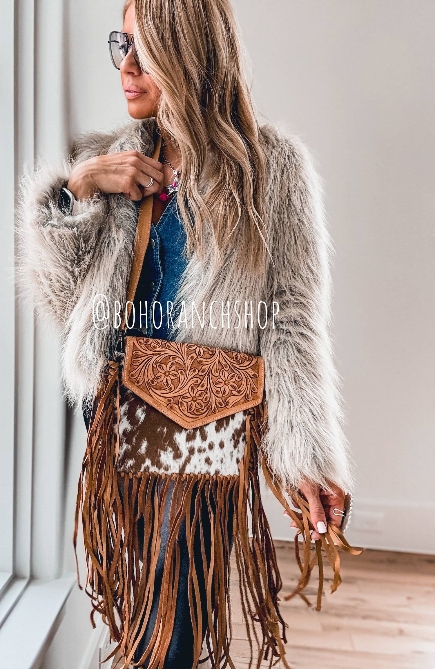 A person wearing a grey coat is holding a brown cowhide crossbody bag with fringe.