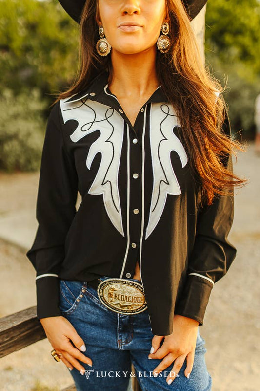 A woman wearing a long sleeve button down shirt with white embroidery yoke, paired with a cowboy hat and jeans.