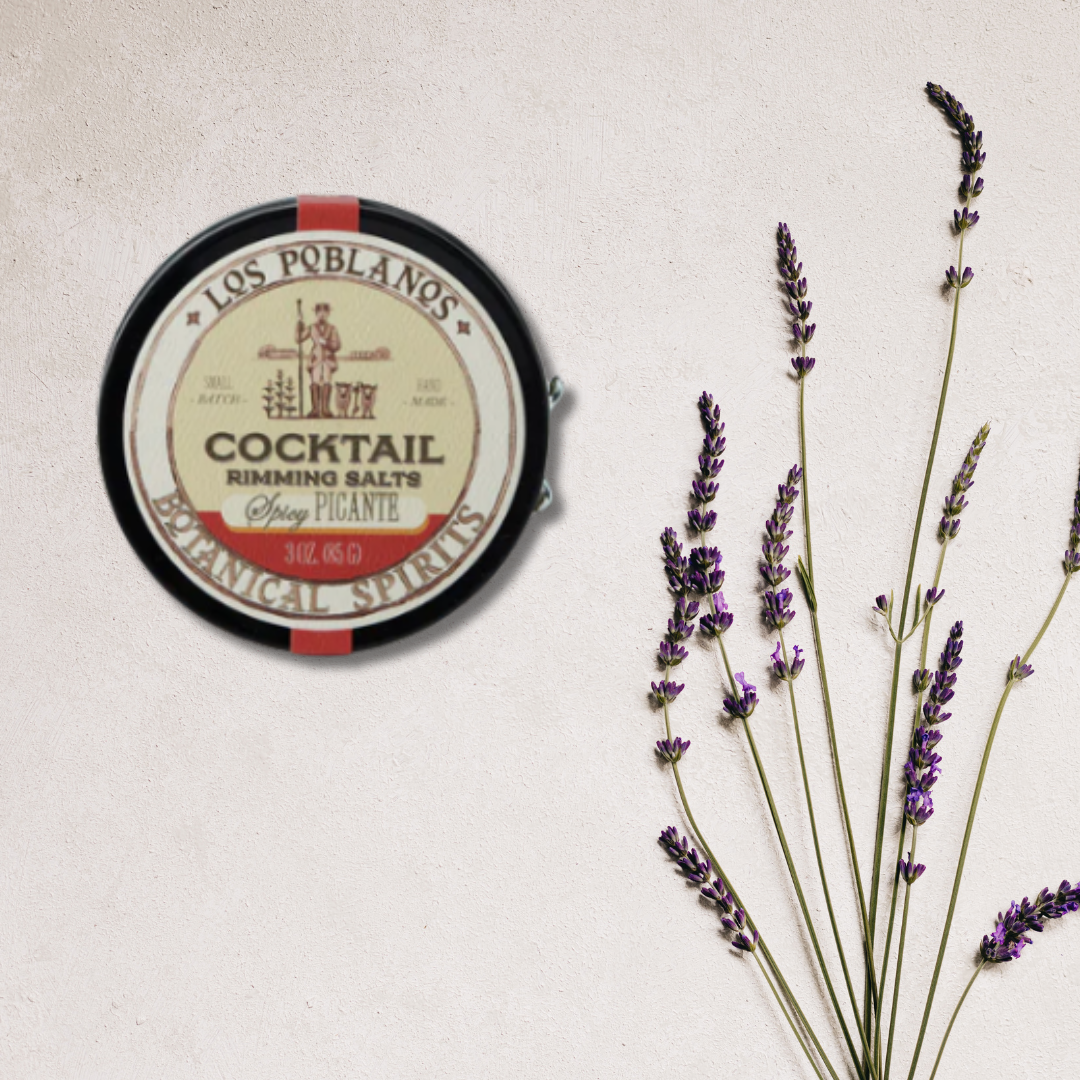 A round tin of Los Poblanos Cocktail Rimming Salt with sea salt flakes next to it, against a white background with some decorative lavender sprigs to the side.