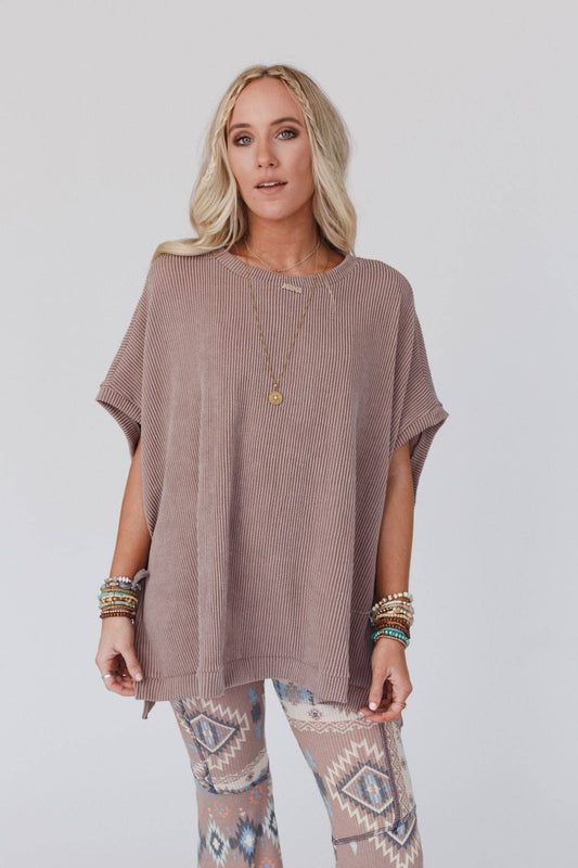 A female model wearing an oversized knit top in brown color, paired with patterned leggings, accessorized with a necklace and multiple bracelets.
