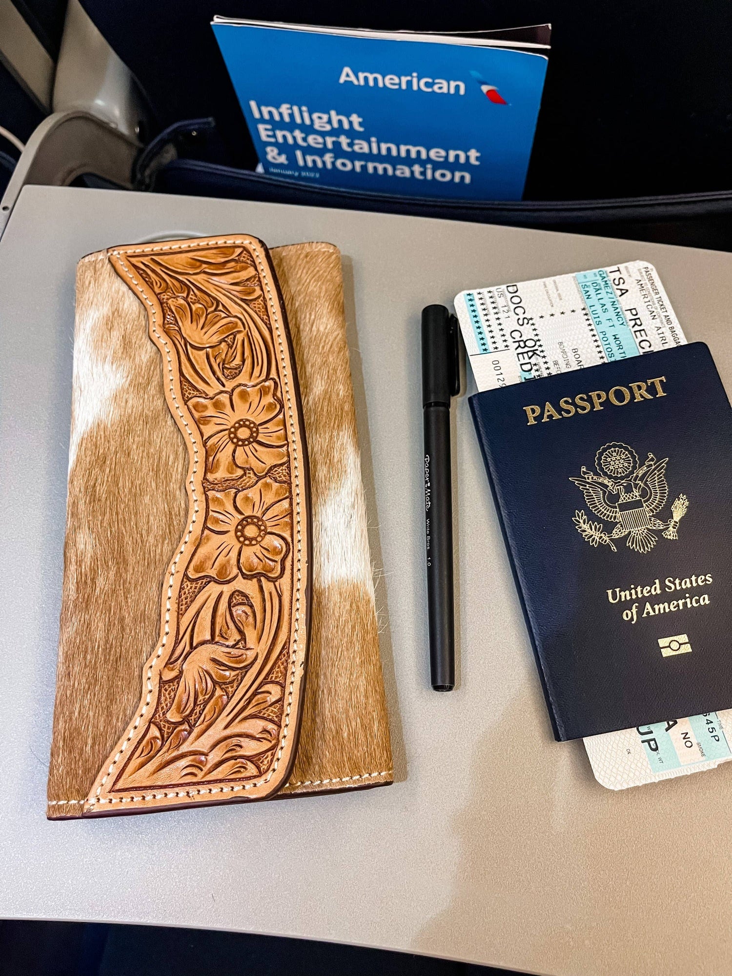 A leather cowhide wallet with intricate tooling, displayed next to a passport, boarding pass, and an inflight entertainment guide on an airplane seat.