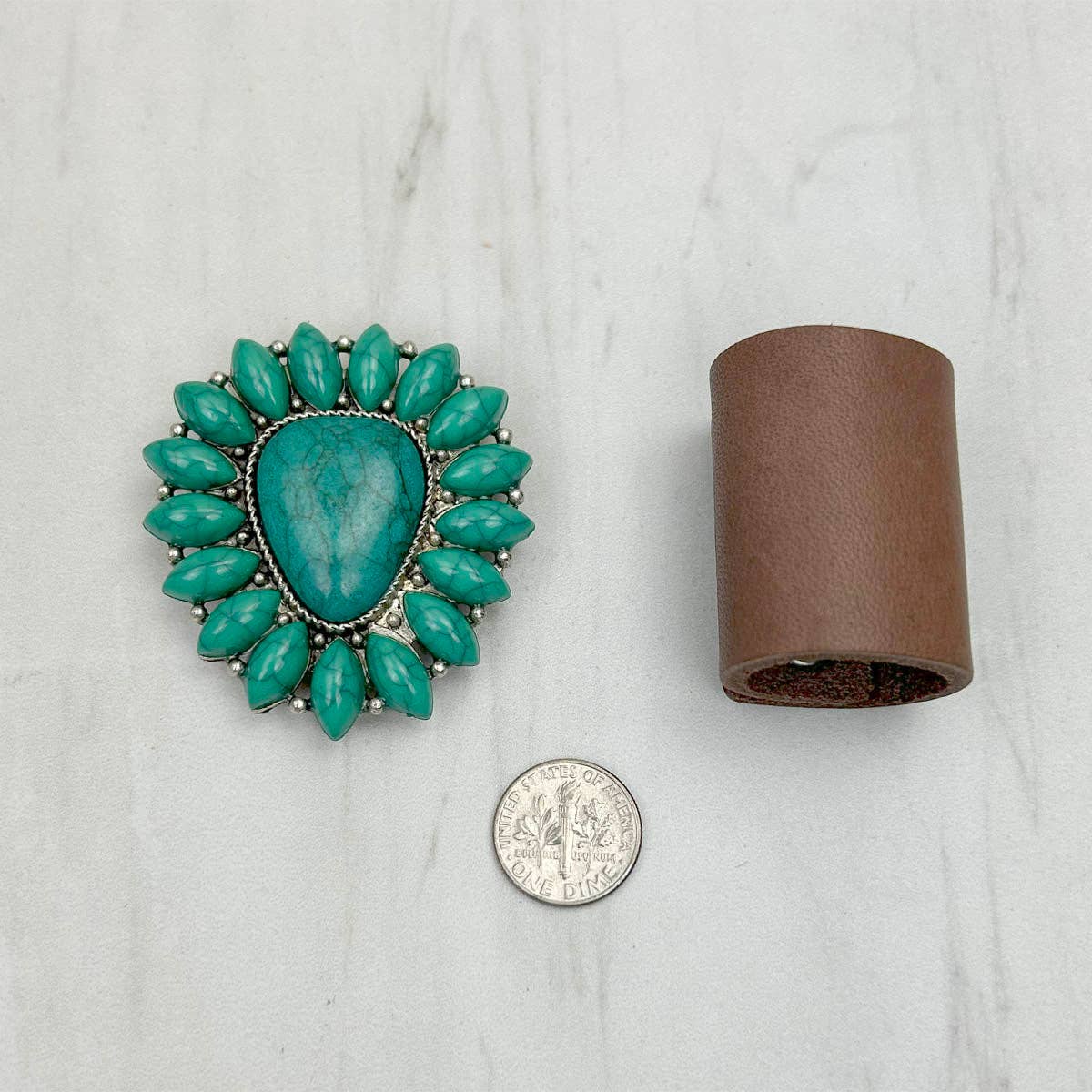 Turquoise brooch next to a cylindrical brown object and a silver coin on a light gray background