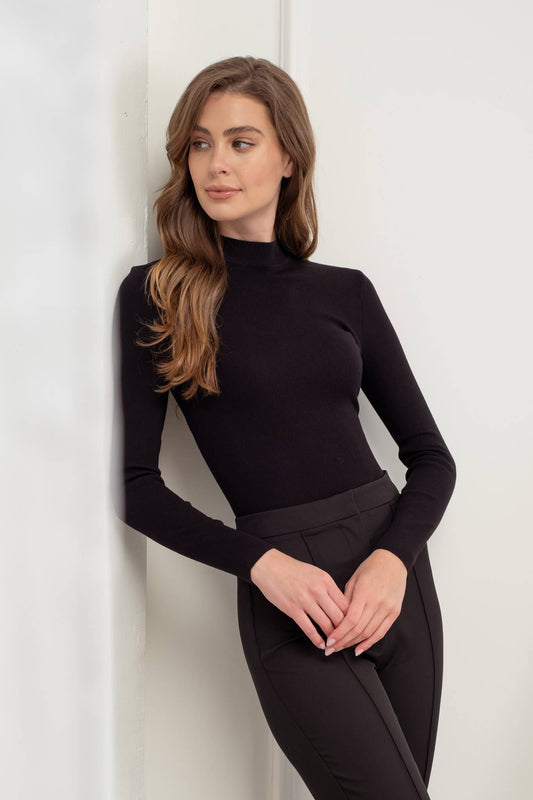 A woman wearing a black mock neck pullover and black pants, standing against a white background.