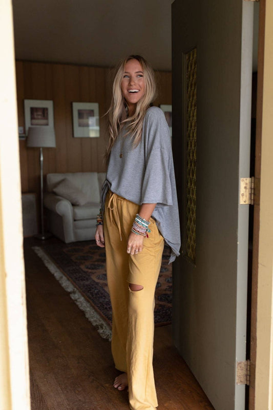 A person wearing an oversized heather grey tee with short sleeves and a ruffled edge on the split back detailing. The person is paired this with mustard yellow wide-leg pants.
