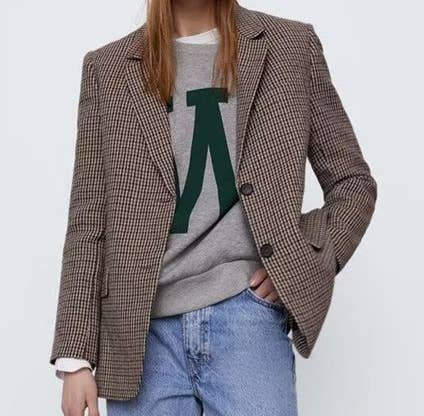 Person wearing a plaid blazer over a gray sweater with a green logo, standing against a plain background.