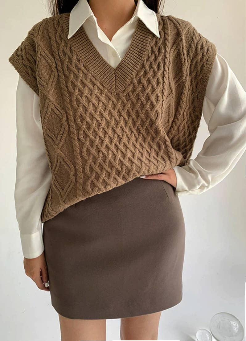 Person wearing a brown knitted vest over a white shirt and brown skirt on a plain background