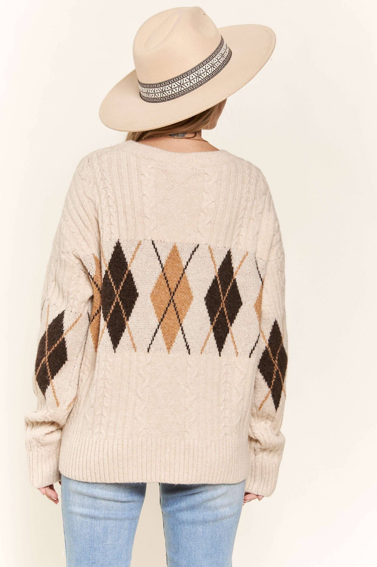 Person wearing a beige sweater with argyle pattern and a hat on a plain background