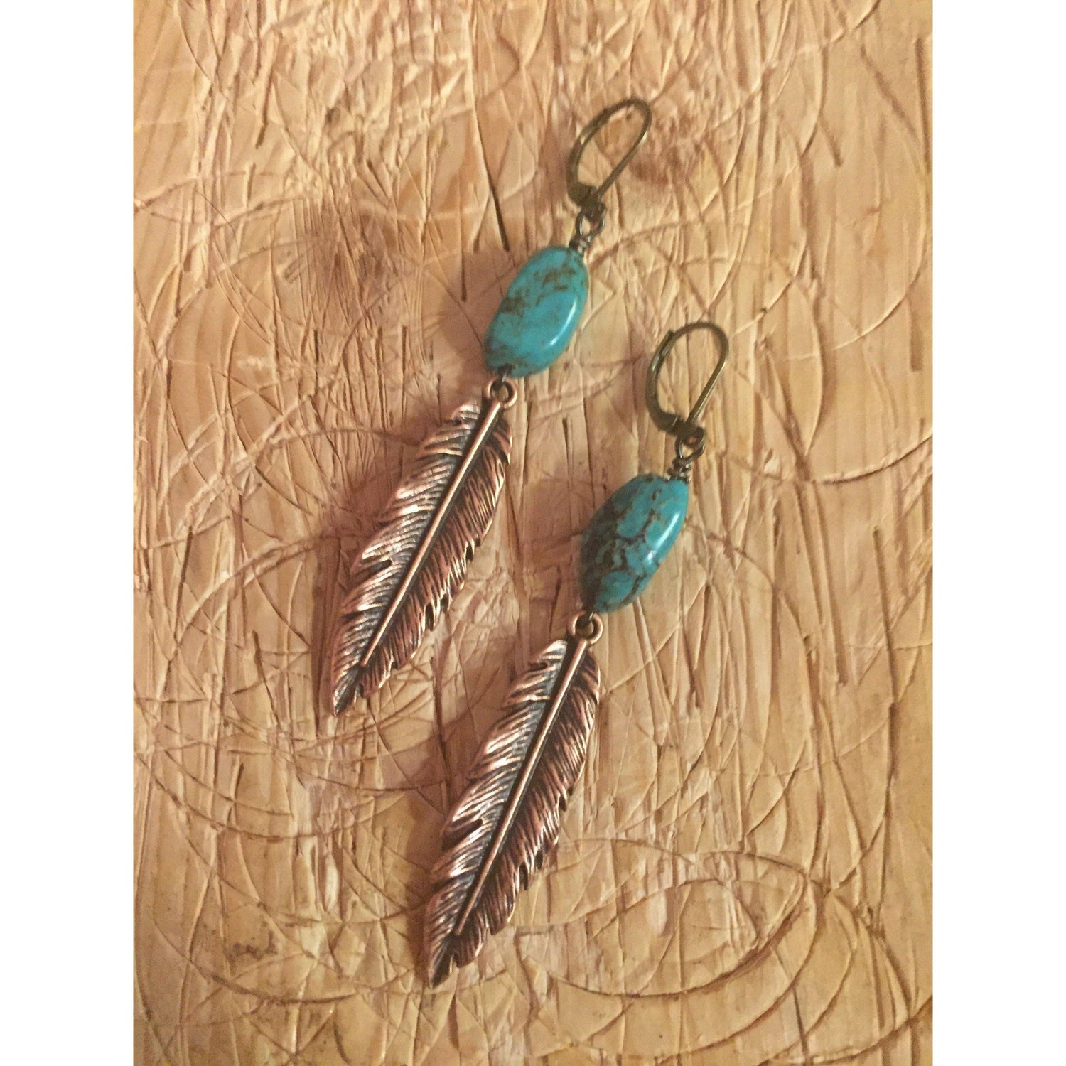 A pair of earrings with copper feather charms and turquoise beads attached to a textured background.