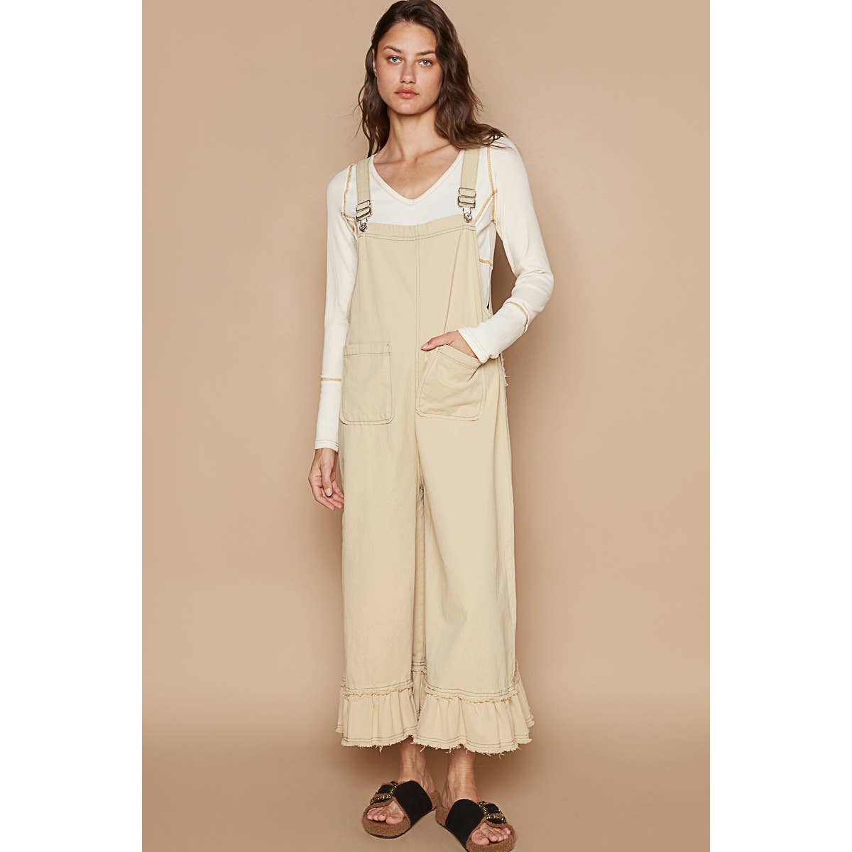 A female model wearing beige overalls with ruffle detail and back pockets, paired with a white top and black sandals, standing against a beige background.