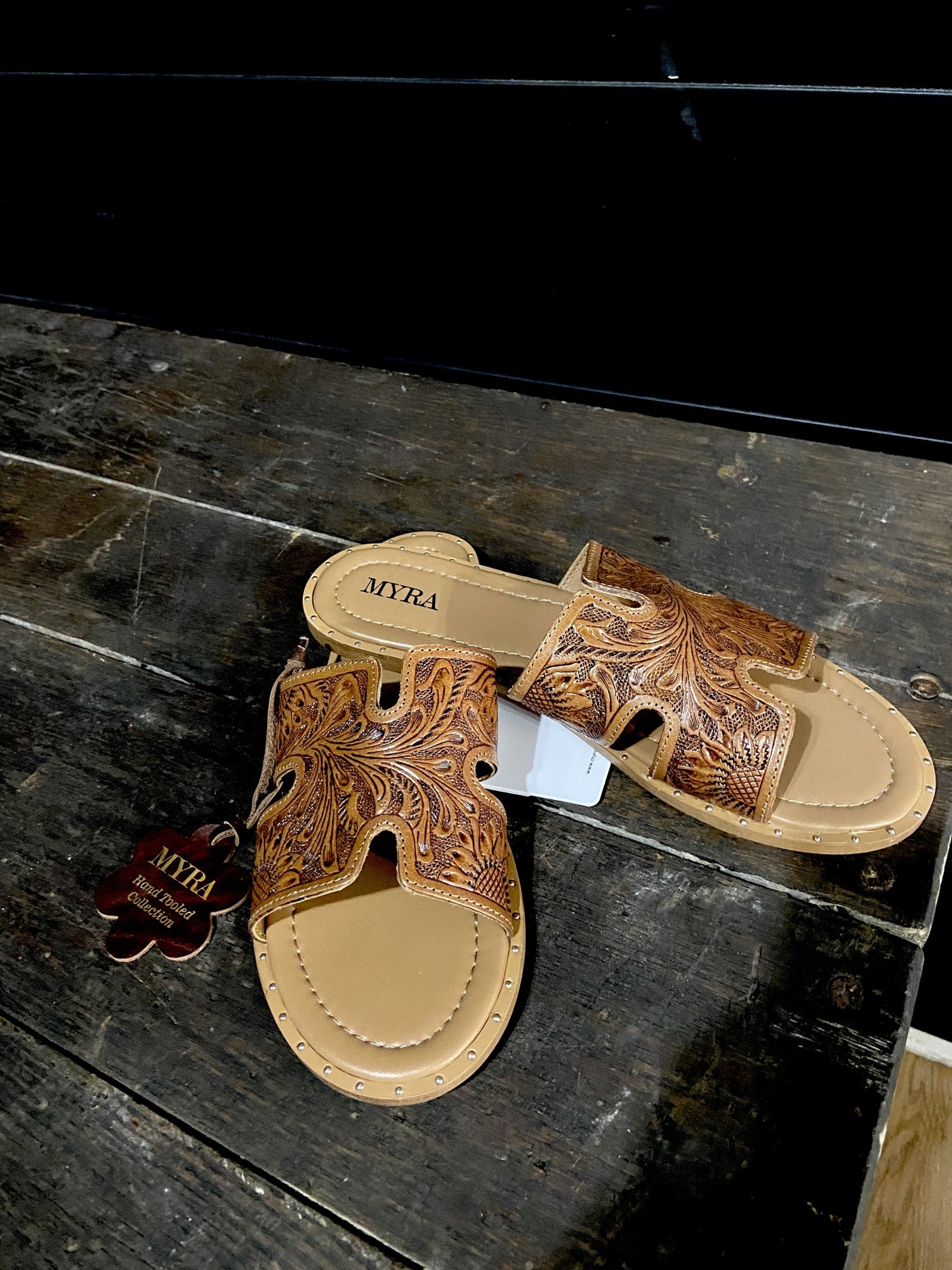 Artisanal leather slides featuring intricate tooled floral designs with rich cognac hue and classic H-strap silhouette blend Western charm with modern sophistication.